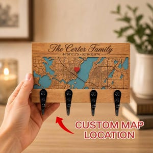 May include: A rectangular wooden wall-mounted key holder featuring a laser-engraved map with blue water details, a small red heart marker, the text The Certer Family, coordinates 44°58'17.2"N 93°16'32.8"W, and four black metal hooks.