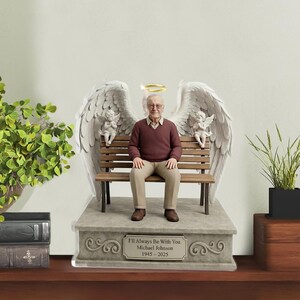 May include: A commemorative figurine featuring a man in a maroon sweater and tan trousers sitting on a wooden bench. Large white angel wings extend behind him, with a gold halo above his head and two small cherubs on the bench. The base reads: I'll Always Be With You. Michael Johnson 1945-2025.