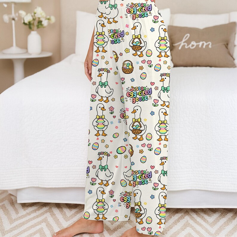 Puede incluir: Pantalones de pijama color crema con un estampado de Pascua. El dise&ntilde;o presenta gansos de dibujos animados sosteniendo huevos y cestas de Pascua, junto con flores y el texto "Just an egg-cited goose".