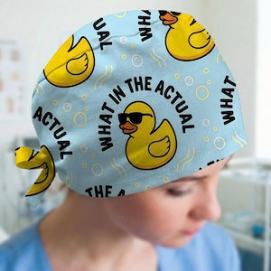 May include: A light blue scrub cap featuring a repeating pattern of yellow rubber ducks wearing black sunglasses. The text WHAT IN THE ACTUAL is printed in black capital letters throughout the design, accompanied by small white circular bubbles.