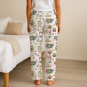 Puede incluir: Pantalones de pijama blancos con un estampado integral de dibujos de gansos sosteniendo huevos y cestas de Pascua, con el texto "Just an egg-cited goose". El dise&ntilde;o incluye huevos y flores en colores pastel.