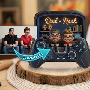 May include: A custom 3D display featuring a black video game controller frame with blue accents. Inside the frame are two cartoon-style figurines of a man and a boy sitting on a couch. The text "Dad - Noah" is displayed in glowing yellow script.
