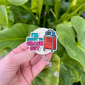 Peut inclure: Un badge blanc avec le texte "I'M ABOUT TO CRASH OUT" en bleu et rose. Le badge présente une image de dessin animé d'un chariot médical rouge avec un moniteur bleu.