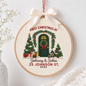 Könnte beinhalten: Ein runder Stickrahmen-Schmuck auf cremefarbenem Stoff mit einer gestickten grünen Tür, zwei Weihnachtsbäumen, Geschenken und Laternen. Der Text lautet: FIRST CHRISTMAS AT Johnny & Sohia 25 JOHNSON ST. 2026. Mit einer weißen Satinschleife.