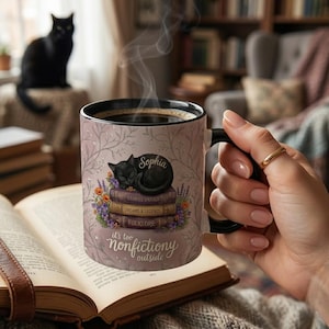 May include: A ceramic mug with a black handle and rim, filled with coffee. The mug features a sleeping black cat on a stack of books with the words "Stories Untold", "Dreams & Legends", and "Folklore". The mug also has the text "it's too nonfictiony outside".