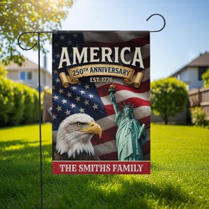 Op de afbeelding: Een tuinvaandel met de Amerikaanse vlag, het Vrijheidsbeeld en een adelaar. De vlag leest "AMERICA 250th ANNIVERSARY EST. 1776" met "THE SMITHS FAMILY" onderaan. De vlag staat op een zwarte metalen standaard in een grasrijke tuin.