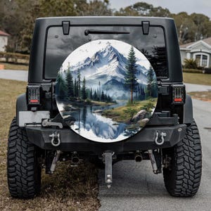 May include: A black Jeep with a spare tire cover featuring a mountain and lake landscape scene. The artwork depicts snow-capped mountains, evergreen trees, and a reflective lake. The tire cover is circular and mounted on the rear of the vehicle.