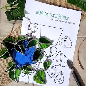 May include: A stained glass hanging plant with green leaves and a blue center. The design is next to a printed pattern sheet with the text "HANGING PLANT PATTERN BY: COLOURCUT STUDIO".