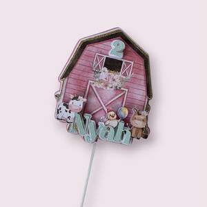 May include: A pink barn-shaped cake topper with a gold trim, featuring a cow, chicken, and horse. The number 2 is at the top, and the name "Nyah" is written in a light blue font. A silver stick supports the topper.
