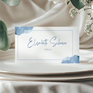 May include: A white place card with a blue watercolor design. The card features the name "Elisabeth Schewe" in a delicate script font, with "TABLE 1" below. Blue watercolor accents frame the top and bottom corners.