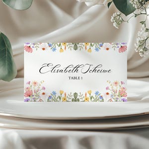 May include: A white place card with floral watercolor border. The card features the name "Elisabeth Scheiwe" in elegant script and "TABLE 1" below. The floral border includes pink, yellow, blue, and purple flowers.