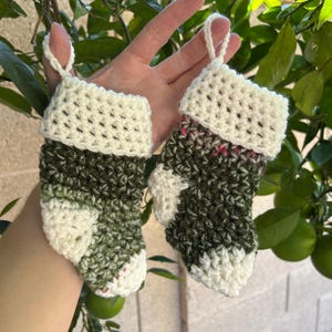 May include: Two miniature crochet Christmas stockings. The stockings are primarily green and white, with a white cuff and toe. Each stocking has a small loop for hanging. The background is a green leafy tree.