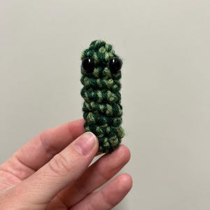 May include: A handmade, green and dark green crocheted creature with two black plastic eyes. The small, cylindrical figure is held up against a neutral background. The yarn is tightly woven, creating a textured appearance.