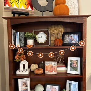 May include: A wooden bookshelf decorated with fall-themed items. Includes a painting of trees, a large letter "G", stacked orange pumpkins, a clock, framed photos, and a garland of crocheted flowers. Various pumpkins and decorative items are displayed.
