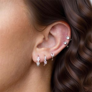 Può includere: Primo piano di un orecchio adornato da molteplici orecchini. Gli orecchini includono piccoli cerchi con disegni a farfalla e floreali, e delicati borchie. I gioielli sono argentati e presentano pietre preziose chiare e rosa, creando un effetto scintillante.