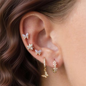 May include: A collection of gold-toned earrings, including butterfly and floral designs, adorn an ear. The earrings feature clear and iridescent stones, creating a delicate and elegant aesthetic. The butterfly earrings dangle, adding movement.
