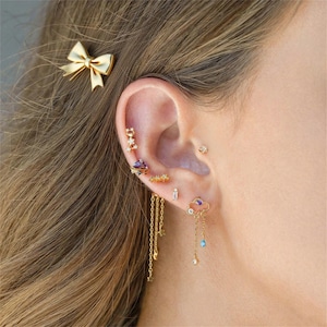 May include: A collection of gold earrings and a bow hair clip adorn an ear. The earrings include studs, hoops, and dangling styles with gemstones and chain details. The bow hair clip is gold with a white center.