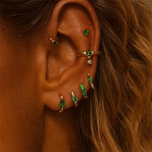 Puede incluir: Primer plano de una oreja adornada con múltiples joyas de oro. La oreja presenta una variedad de pendientes, incluyendo tachuelas, aros y un anillo, todos adornados con piedras preciosas verdes y pequeños diamantes. Las joyas se destacan sobre el fondo de cabello ondulado, castaño claro.