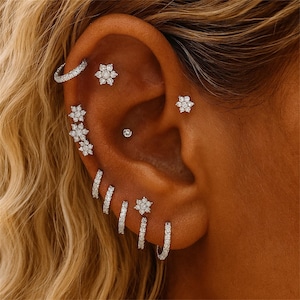May include: Close-up of an ear adorned with multiple pieces of jewelry. The ear features a variety of earrings, including small hoops, studs, and floral designs, all sparkling with what appears to be clear gemstones. The jewelry is set against the backdrop of blonde hair.