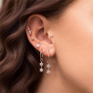Puede incluir: Una oreja adornada con múltiples joyas. Hay varios pendientes pequeños, incluyendo un melocotón rosa, una flor de cuatro pétalos y un grupo de pequeños cristales. Dos pendientes de aro dorados cuelgan con tres colgantes de flores de cristal.