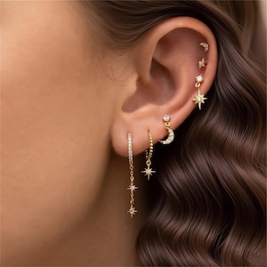 May include: A selection of gold earrings, including hoops, studs, and drop designs. The earrings feature celestial motifs such as stars and crescent moons, embellished with small, sparkling gemstones. The jewelry is displayed on an ear, showcasing the various styles.