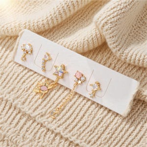 May include: A collection of five gold-coloured earrings presented on a white card. The earrings showcase diverse designs, such as stars, moons, and geometric forms, embellished with clear and pink gemstones. The earrings are set against a cream-coloured knitted background.