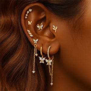 Puede incluir: Una variedad de pendientes dorados, incluyendo diseños de mariposas y hojas, adornan una oreja. Algunos pendientes presentan piedras preciosas transparentes. La colección incluye pendientes de botón, aros y estilos colgantes, creando un look elegante.