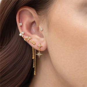 Op de afbeelding: Een oor is versierd met een verzameling goudkleurige oorbellen. De oorbellen omvatten kleine studs, bloemmotieven en bungelende stervormige oorbellen met kettingaccenten. De oorbellen zijn versierd met kleine, sprankelende steentjes.