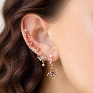 May include: A selection of gold-coloured earrings, featuring celestial and heart designs. The earrings include a planet-shaped dangle earring with a pink heart-shaped gem, and other studs with pink and clear gems. The earrings are displayed on an ear.