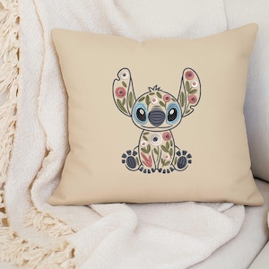 May include: Beige square cushion with an embroidered floral Stitch design. The design features blue, pink, and white flowers, with blue eyes and a black nose. The cushion is placed on a white blanket.