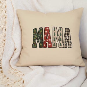 Puede incluir: Un cojín cuadrado beige con la palabra "MAMA" en un diseño festivo. Las letras están decoradas con motivos navideños: acebo, estrellas, árboles de Navidad y cuadros. El cojín está sobre una manta blanca texturizada.