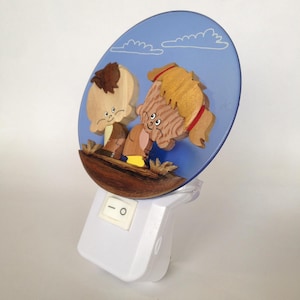 Op de afbeelding: Een grillig nachtlampje met een houten reliëf van twee stripfiguren met lichtbruin en blond haar, tegen een blauwe achtergrond met witte wolken. Het nachtlampje heeft een witte basis met een aan/uit-schakelaar.