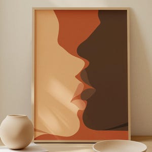 May include: A framed art print featuring two profiles in shades of orange and brown, suggesting a kiss. The artwork is displayed on a light wooden table with a beige vase, books, and a bowl. The room has neutral-toned walls.