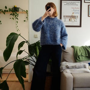 Puede incluir: Un suéter azul de punto difuso con cuello redondo y mangas largas. El suéter se combina con pantalones azul oscuro. La imagen está tomada en interiores, con un sofá y plantas en el fondo. El suéter parece estar hecho a mano.