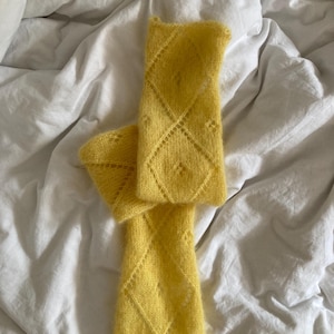Może przedstawiać: Para jasnożółtych dzianinowych ocieplaczy na ramiona z rombowym wzorem. Ocieplacze na ramiona wykonane są z miękkiej, puszystej przędzy i leżą na białej prześcieradle. Rombowy wzór dodaje elegancji projektowi.