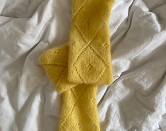 May include: A pair of bright yellow knitted arm warmers with a diamond pattern. The arm warmers are made of a soft, fuzzy yarn and are laying on a white sheet. The diamond pattern adds a touch of elegance to the design.