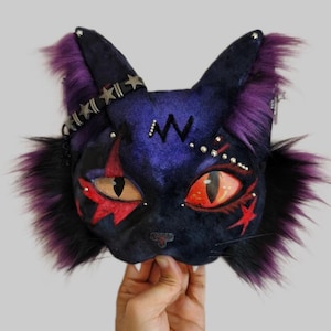 May include: A plush cat head decoration featuring deep purple velvet fabric, vibrant orange eyes, and long purple and black faux fur accents. The face is adorned with embroidered red star shapes, a zigzag pattern, and a black strap with silver stars.