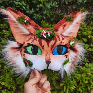 May include: A handmade cat mask with a brown and white spotted pattern, green and blue eyes, and faux fur accents. The mask is decorated with moss and miniature mushroom details.