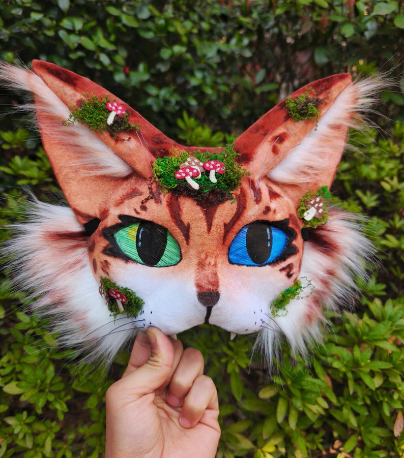 May include: A handmade cat mask with a brown and white spotted pattern, green and blue eyes, and faux fur accents. The mask is decorated with moss and miniature mushroom details.