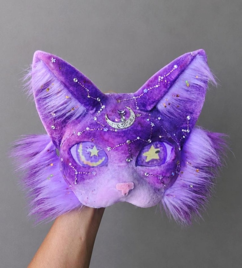 May include: A plush cat mask in shades of purple and lavender, featuring long faux fur on the sides, a silver crescent moon charm on the forehead, and scattered rhinestones forming constellation patterns. The eyes display star and moon motifs.