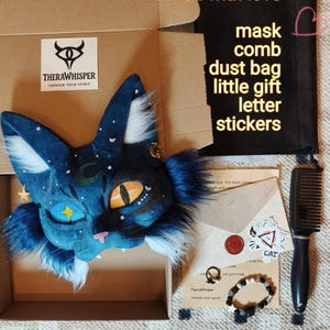 May include: A blue cat mask with star and moon details, inside a cardboard box. The box also contains a black comb, dust bag, gift, letter, and stickers. The text "Packed with love" is visible.