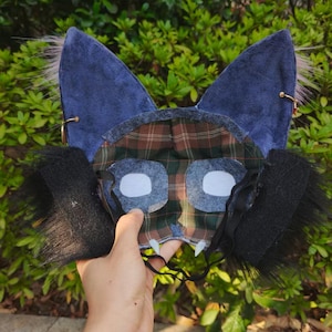 May include: A mask featuring large, pointed blue ears with tufts of fur, a green and brown plaid fabric face, two white eye cutouts, black fur side panels, and two small white fangs at the bottom edge, held by a thin black elastic strap.