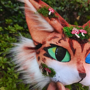May include: A close-up of a cat mask with orange and white fur, green eyes, and small red and white mushroom accents. The mask has a realistic design with a black nose and whiskers, set against a green leafy background.
