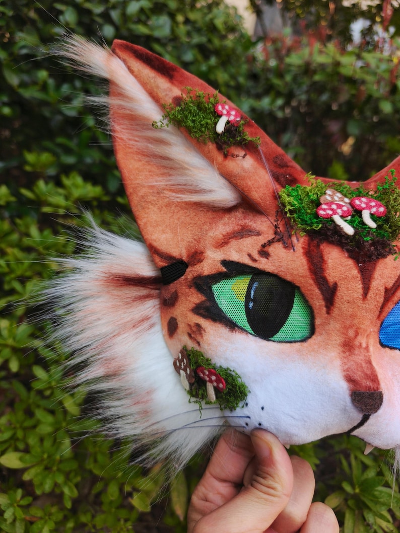 May include: A close-up of a cat mask with orange and white fur, green eyes, and small red and white mushroom accents. The mask has a realistic design with a black nose and whiskers, set against a green leafy background.