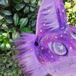 May include: A purple plush animal mask featuring long faux fur, a mesh eye covering with a yellow crescent moon and star graphic, and decorative clear rhinestones arranged in constellations across the velvet-textured surface.