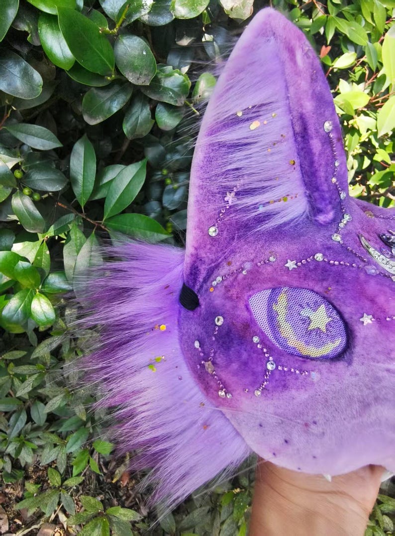 May include: A purple plush animal mask featuring long faux fur, a mesh eye covering with a yellow crescent moon and star graphic, and decorative clear rhinestones arranged in constellations across the velvet-textured surface.