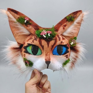 Puede incluir: Una máscara de gato con pelaje marrón y naranja, ojos verdes y azules y bigotes blancos. La máscara está decorada con musgo verde y pequeños acentos de hongos rojos y blancos. Las orejas son largas y puntiagudas, con ribete de piel blanca.