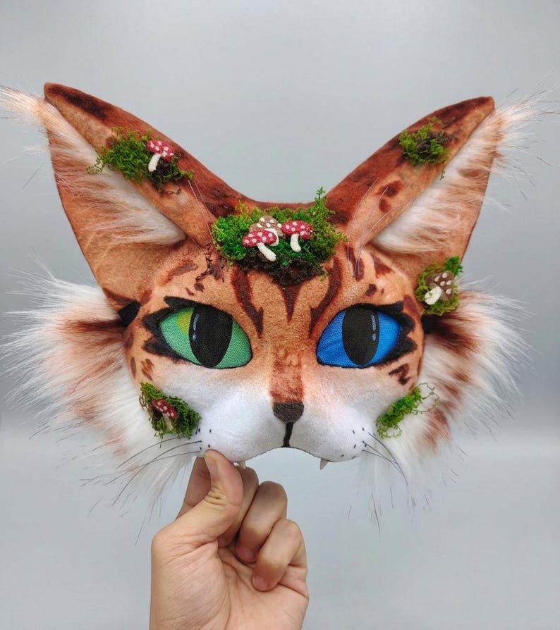 May include: A cat mask with brown and orange fur, green and blue eyes, and white whiskers. The mask is decorated with green moss and small red and white mushroom accents. The ears are long and pointed, with white fur trim.