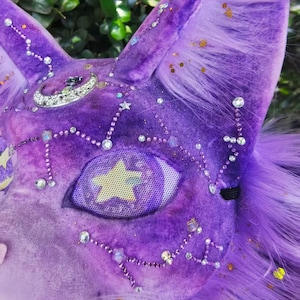 May include: A purple plush animal mask featuring long faux fur, a silver crescent moon charm, and scattered clear rhinestones forming constellation patterns. The eye area is covered with a mesh screen displaying a yellow star graphic.