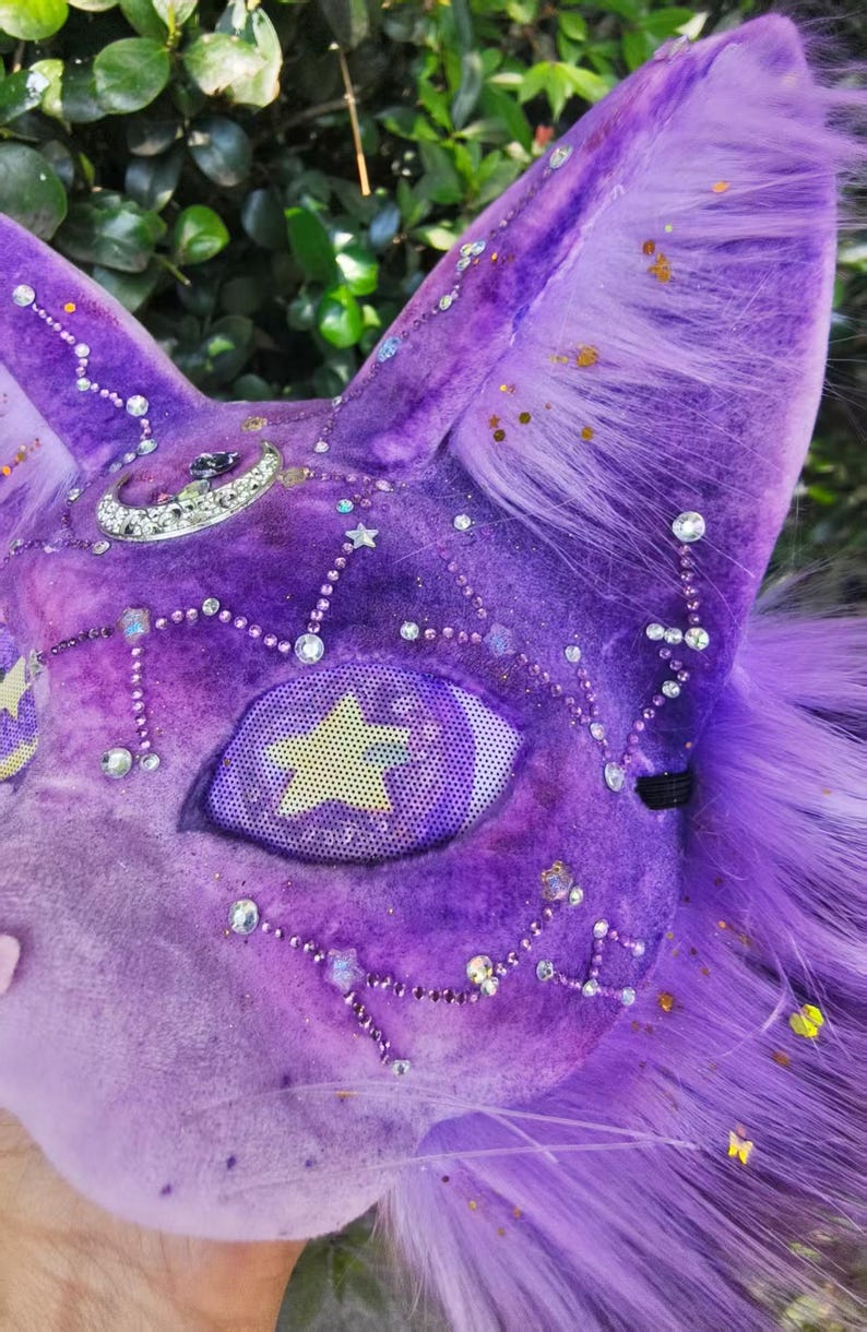 May include: A purple plush animal mask featuring long faux fur, a silver crescent moon charm, and scattered clear rhinestones forming constellation patterns. The eye area is covered with a mesh screen displaying a yellow star graphic.